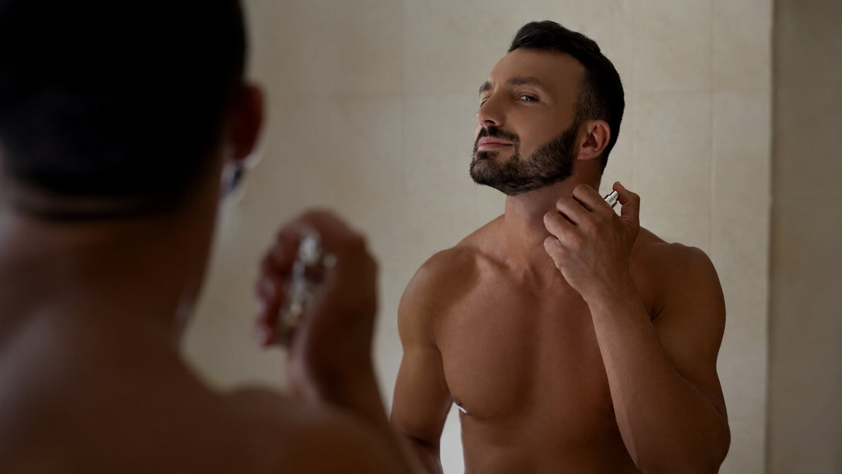 A shirtless man with a beard applies a fragrance from a silver atomizer mirror in a bathroom.