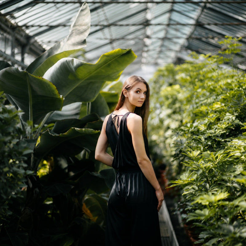 A woman in a sleeveless black dress stands in a lush greenhouse surrounded by large tropical plants under a glass roof.