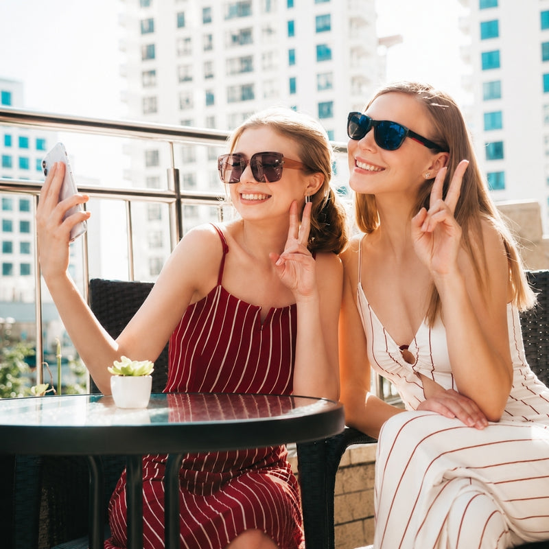 Two women posing on a sunny balcony with a glass table, taking a selfie. One wears a red and white striped sleeveless dress and sunglasses, while the other wears a white and red striped dress and dark sunglasses.
